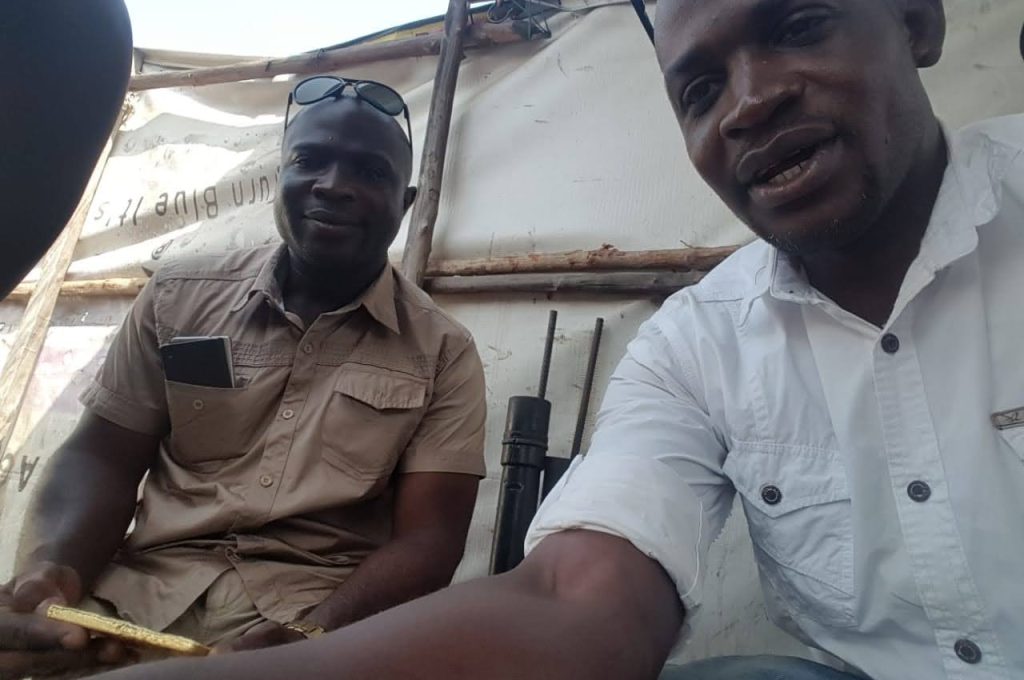 Inside a tent, two men are seated, with one man holding a gold block, symbolizing employees of the new Liberian mining company.
