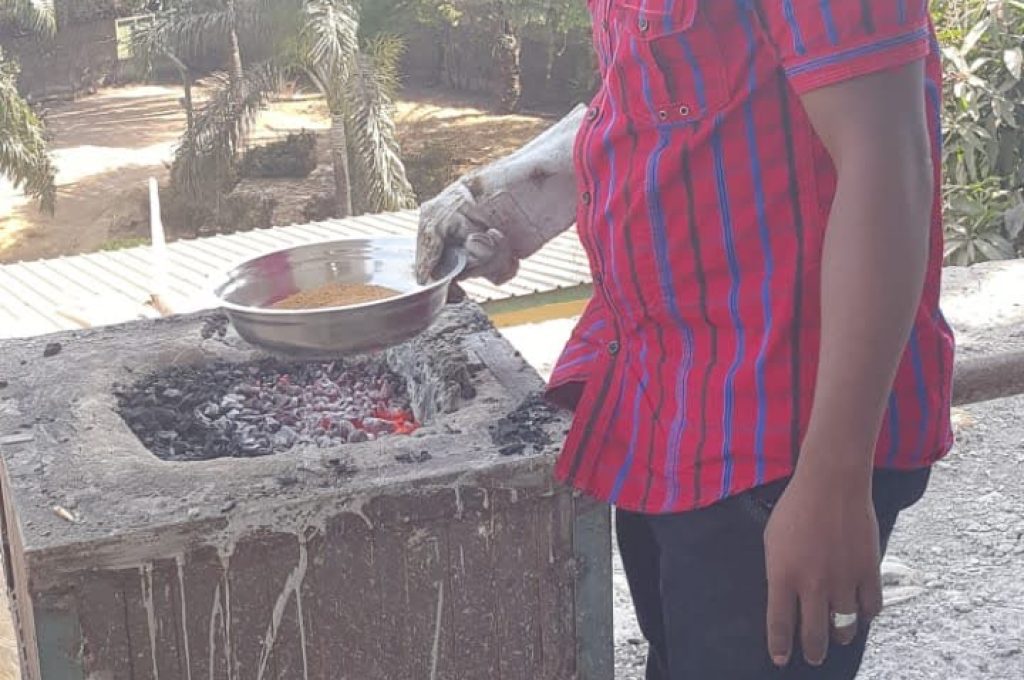 A man in a red and blue shirt put a pan fill with gold dust on an outdoor grill at the New Liberian Mining Company site, with mining work in progress.